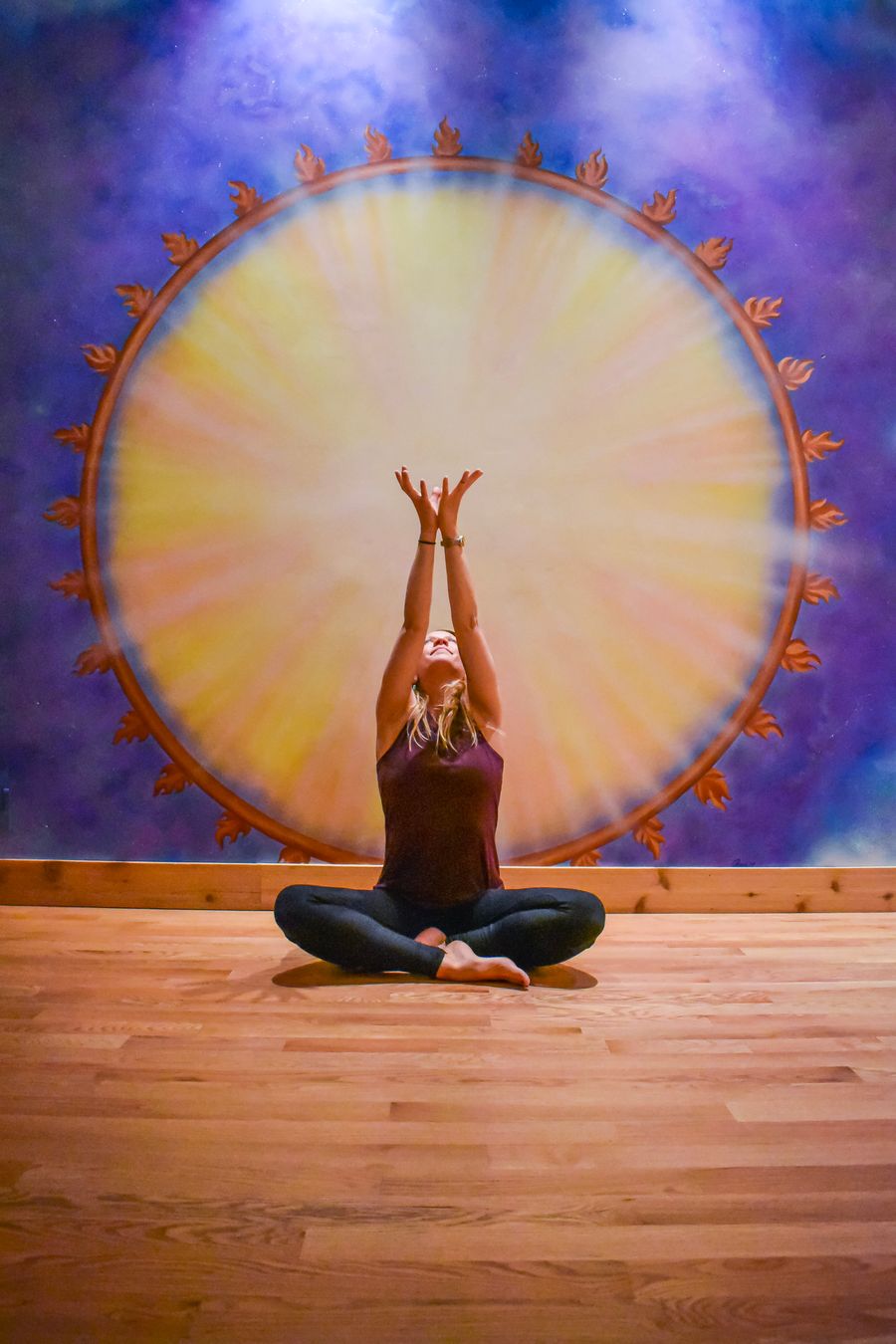 Seated cross-legged with the arms raised, framed by the sun mural